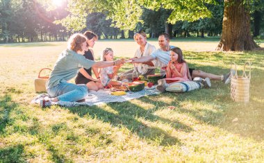 Hafta sonu güneşli bir günde şehir parkında piknik battaniyesinde oturan büyük bir aile. Gülüyorlar, gülüyorlar ve karpuz yiyorlar, haşlanmış mısır, soğuk çayla turta yiyorlar. Aile değerleri, açık hava aktiviteleri konsepti.