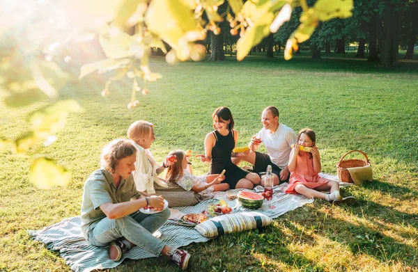 周末阳光明媚的一天 一家人坐在城市公园里的野餐毯上 他们笑着 笑着吃着煮熟的玉米和西瓜 家庭价值观和户外活动概念 — 图库照片