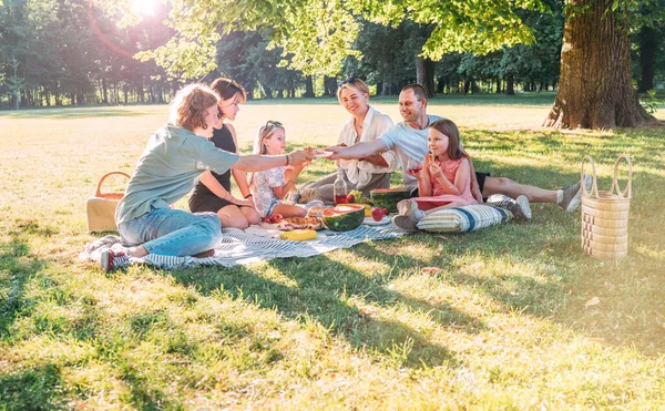 Big family sitting on the picnic blanket in city park during weekend sunny day. They smiling, laughing and eating watermelon, boiled corn,pie with cold tea. Family values, outdoors activities concept.