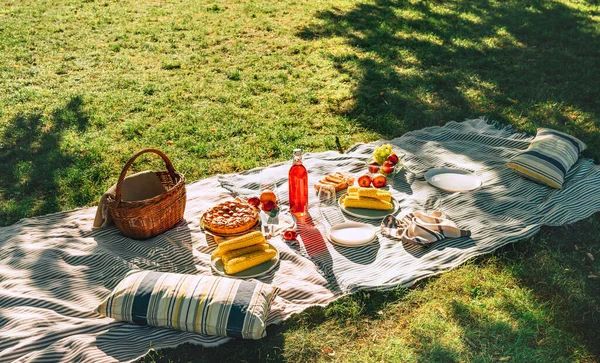 Picnic blanket with boiled corn, fruits, just backed pie, a bottle of juice, basket still life in the city summer park. No people.