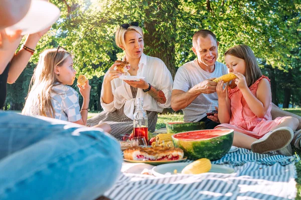 日曜日の晴れた日には 市内の公園のピクニックブランケットに座っている大きな家族 彼らは笑顔で 笑って ゆでたトウモロコシとスイカを食べている 家族の価値観とアウトドア活動のコンセプト — ストック写真