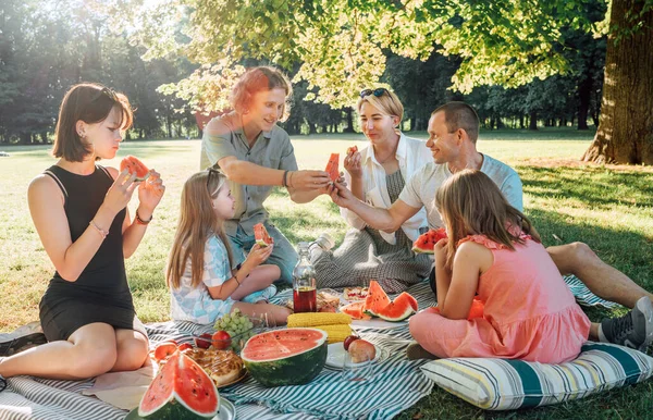 Big family sitting on the picnic blanket in a city park during weekend Sunday sunny day. They are smiling, chatting, laughing and eating juicy watermelon. Family values and outdoor activities concept.