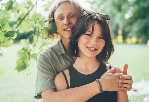 若い10代の男の子は 夏の都市公園での屋外散歩中にカメラに笑顔で彼のガールフレンドを抱きかかえています 最初の愛 現代のティーンエイジャーの関係の概念イメージ — ストック写真