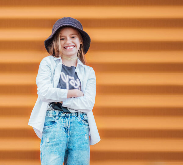 Маленький модный женский портрет искренне улыбается, глядя на камеру на фоне оранжевой стены. Городские жители и образ концепции уличной повседневной жизни 