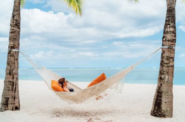 Young woman relaxing in wicker hammock on the sandy beach on Mauritius coast and enjoying wide ocean view waves. Exotic countries vacation and mental health concept image.