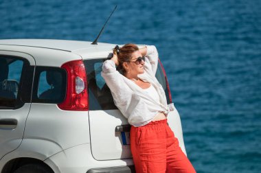 Güneş gözlüklü yalnız bir kadın, yol kenarındaki küçük beyaz arabanın yanında açık yaz kıyafetleri içinde Yunanistan 'daki Cephalonia adasında İyon deniz manzarasının tadını çıkarıyordu. Yaz tatili ya da otomobil seyahat konsepti