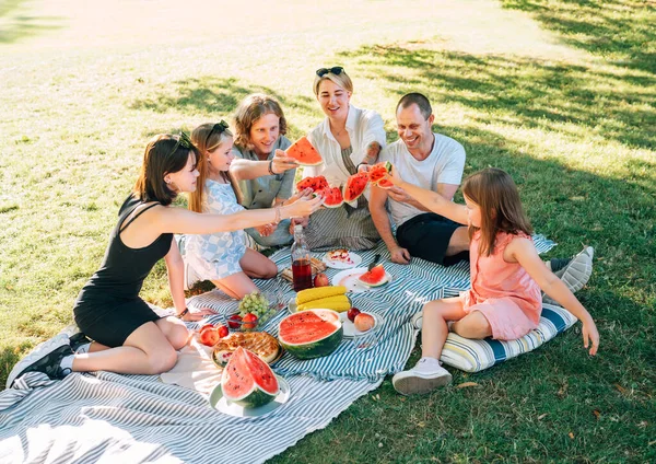 Big caucasian family on picnic blanket on the in city park green grass. They rose-up red juicy watermelon pieces and clinking them like glasses. Family values and outdoor activities concept.