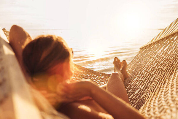 Young woman relaxing in wicker hammock on the sandy beach on Mauritius coast and enjoying sunset light over Indian ocean waves. Exotic countries vacation and mental health concept image. 