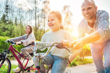 Açık havada yürürken iki kızıyla gülümseyen bir baba. O, genç kıza bisiklet sürmeyi öğretiyor. Onlar yaz şehir parkında birliktelikten hoşlanırlar. Mutlu çocukluk konsepti.