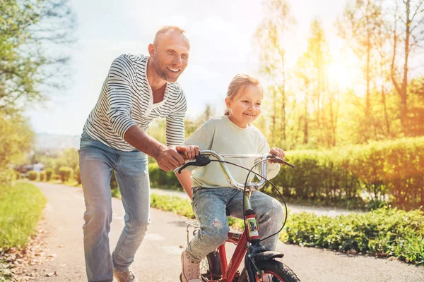 笑顔の父は自転車に乗るように娘に教える 彼らは夏の都市公園で一緒に楽しむ 幸せな子供時代のコンセプトイメージ — ストック写真