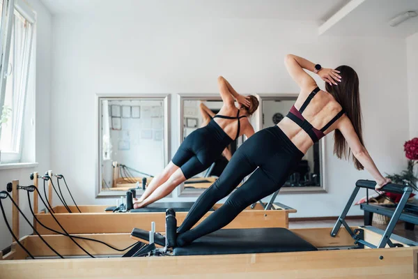 两名面带微笑 体形合适的女性在带镜子的体育运动馆使用普拉提改革机进行侧板运动 提高手臂静心肌肉运动 积极的人员培训概念 — 图库照片