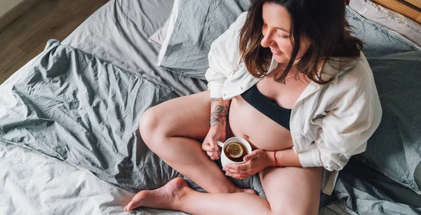 若い妊婦のクロスに座って ベッドルームでお茶のカップとベッドの上に早朝の時間に足 女性の健康 幸せな妊娠と穏やかな精神的な気分の概念イメージ — ストック写真