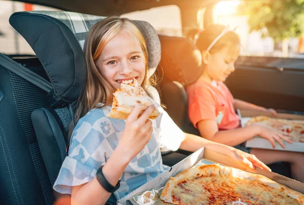 Portrait of positive smiling girl eating just cooked italian pizza sitting with a sister on car back seat in child car seats. Happy childhood, fastfood eating or auto jorney lunch break concept image.