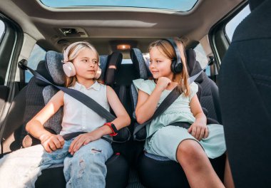 İki pozitif gülümseyen kız kardeşin portresi çocuk koltuklarında oturmuş emniyet kemerlerini bağlayıp kulaklıkla müzik dinliyorlar. Mutlu çocukluk, otomobil yolculuğu konsept resmi.