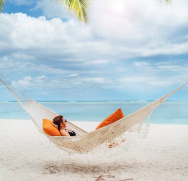 Mauritius sahilindeki kumlu beyaz plajda hasır hamakta dinlenen ve geniş okyanus manzaralı dalgaların tadını çıkaran genç bir kadın. Egzotik ülkeler tatil ve ruh sağlığı konsepti imajı.