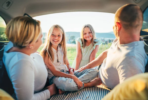 Portrait of happy smiling little sisters. Happy young couple with two daughters inside the car trunk during auto trop. They are smiling, laughing and chatting. Family values, traveling concept.