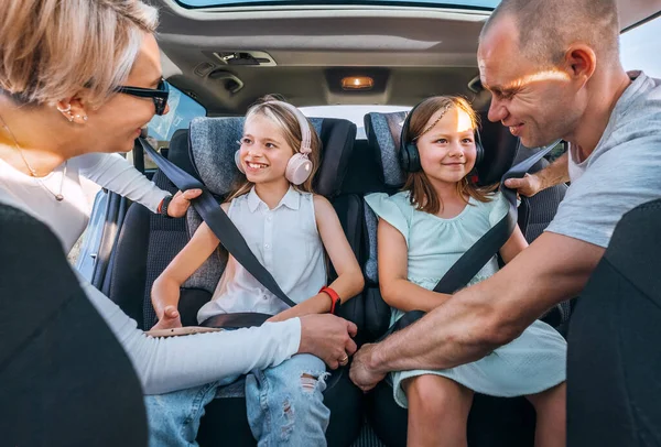 Inside car photo of mother and father fastening with safety auto belts their little daughters girl sitting in child seat and listening headphones. Family values, traveling and technology concept.