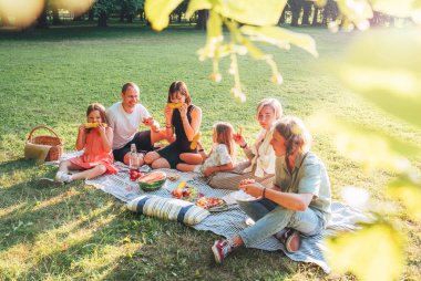Hafta sonu güneşli bir günde şehir parkında piknik battaniyesinde oturan büyük bir aile. Gülümsüyorlar, gülüyorlar ve haşlanmış mısır ve karpuz yiyorlar. Aile değerleri ve açık hava aktiviteleri kavramı.
