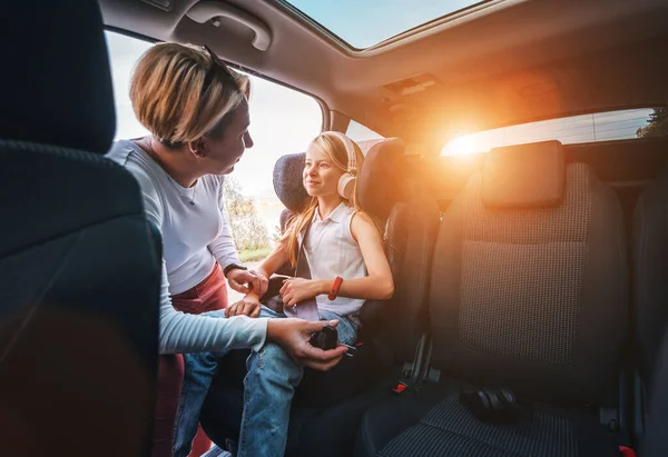 Inside the car photo of a mother fastening with safety auto belt her little daughter girl sitting in child seat. Girl listening music using headphones. Family values, traveling and technology concept