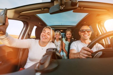 Otomobil kazasında iki kızı olan mutlu genç bir çift. Gülümsüyorlar, gülüyorlar ve akıllı telefon kullanarak selfie çekiyorlar. Aile değerleri, seyahat, sosyal medya ve yeni teknoloji kavramları.