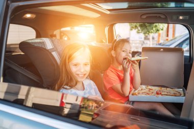 İki pozitif gülümseyen kız kardeş, arabanın arka koltuğunda çocuk koltuklarında pişmiş İtalyan pizzası yemekten mutlu olurlar. Mutlu çocukluk, fast food yemek, ya da otomobil yolculuğu öğle yemeği tatili konsepti.