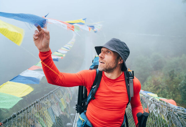 Young smiling man crossing canyon over Suspension Bridge and touching multicolored Tibetan Prayer flags hinged over gorge. Mera peak climbing route trek near Lukla, Sagarmatha National Park, Nepal.