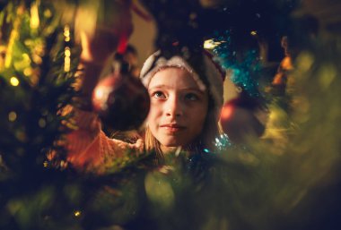 Kırmızı Noel Baba şapkalı şirin küçük kız sihirli ağaç süsleme topunu peri masalı X-Masalı dilek gecesinde Noel ağacına asıyor. Dalların içindeki büyüleyici çocukluk ve kış tatili konsepti