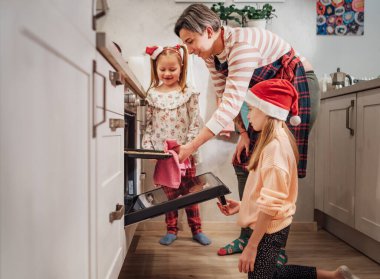 Cute little girls in red Santa hats with mother making homemade sweet biscuits. They putting baking sheet in hot oven. Christmas cookies preparation. Home sweet home warm family life moments concept.	