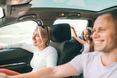 Happy young couple with daughter inside the modern car with panoramic roof during auto trop. They are smiling, laughing during road trip. Family values, traveling concepts.	