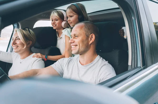 Happy young couple with two daughters inside the modern car with panoramic roof during auto trop. They are smiling, laughing during road trip. Family values, traveling concepts.