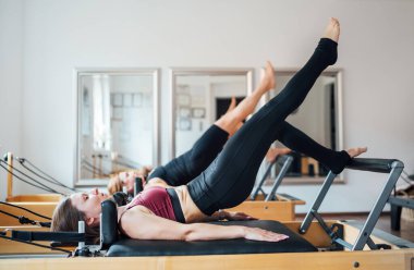 Spor spor salonunda ayna duvarlı pilates reformatörünü kullanarak yükseltilmiş bacak statik kaslarıyla köprü egzersizi yapan formda iki kadın. Etkin insanlar eğitim kavramı