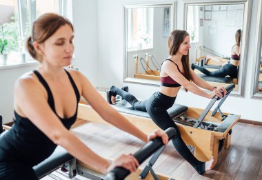 Spor spor salonunda ayna duvarlı, pilates reformatörünü kullanarak esneme egzersizleri yapan gülümseyen iki kadın. Etkin insanlar eğitim veya tıbbi rehabilitasyon konsepti imajı.