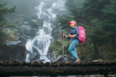 Young woman with backpack and trekking poles crossing wooden bridge near power mountain river waterfall during Makalu Barun National Park trek in Nepal. Mountain hiking and active people concept image clipart