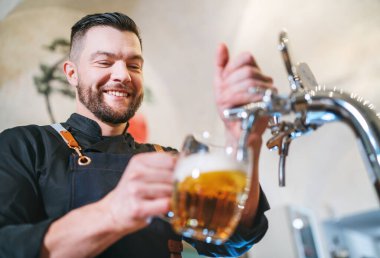 Gülümseyen şık sakallı barmen, siyah üniformalı, önlüklü barmen bardaki bardakta yeni bira içiyor. Başarılı insanlar, bira tüketimi, içecek endüstrisi imajı