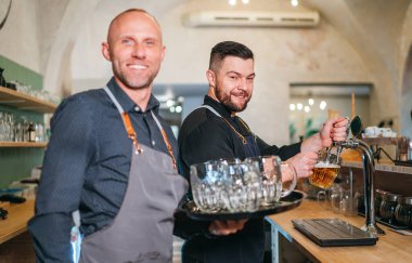 Tarz sahibi sakallı barmen siyah üniformalı barmen bar tezgahına tıklatıyor ve garsonla sohbet ediyor. Birbirlerine gülümsüyor.