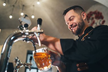 Gülümseyen şık sakallı barmen siyah üniformalı önlüklü barmen bardaki cam bardağa bira tıklatıyor. Başarılı insanlar, bira tüketimi, içecek endüstrisi imajı