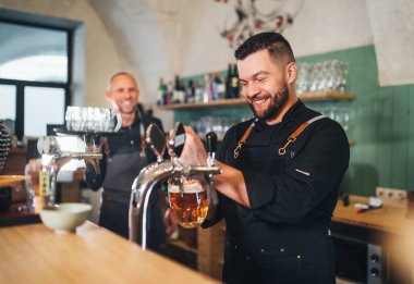 Siyah üniformalı şık sakallı barmen kameraya gülümsüyor, bar tezgahına bira vuruyor ve garsonu tepsiyle dürtüyor. Başarılı insanlar takım çalışması, arkadaşlık, demleme ve restoran endüstrisi konsepti imajı