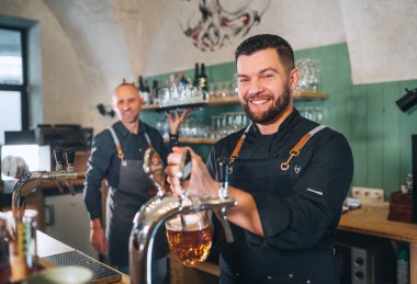 Siyah üniformalı şık sakallı barmen kameraya gülümsüyor, bar tezgahına bira vuruyor ve garsonu tepsiyle dürtüyor. Başarılı insanlar takım çalışması, arkadaşlık, demleme ve restoran endüstrisi konsepti imajı