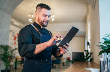 Küçük işletme sahibi, siyah bir şef üniforması içinde, rahat restoranında dijital tablet kullanarak gülümsüyor. Başarılı insanlar, sıkı çalışma, tüketici kafeleri, restoran endüstrisi konsepti.