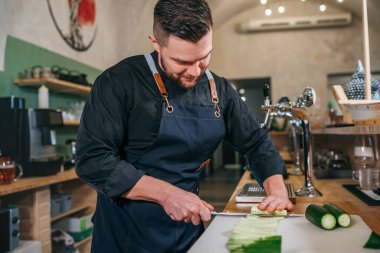 Bearded chef cook dressed black uniform with an apron chopiing fresh green cucumber with sharp knife in basement restaurant kitchen. Professional occupation, food preparation and restaurants industry. clipart