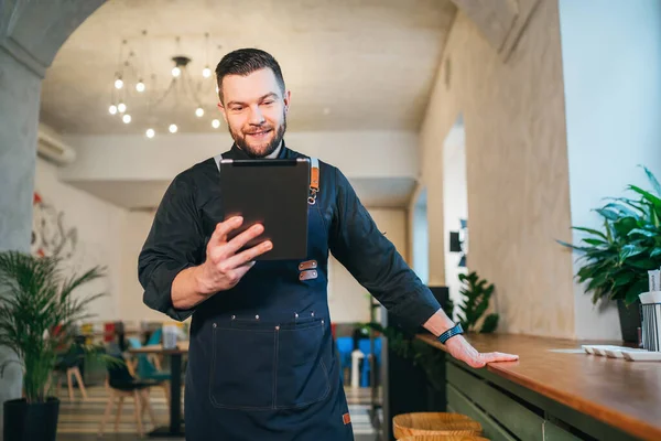 Smiling small business owner dressed in a black chef uniform with an apron using digital tablet in his cozy restaurant hall. Successful people, hard work, consumer cafes, restaurants industry concept.