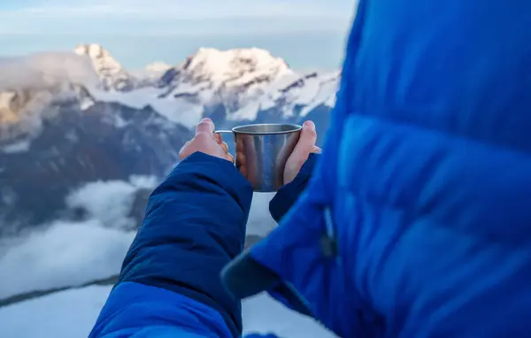 High altitude female mountaineer dressed blue warm dawn jacket holding metal mug of hot tea and drinking warm drink with Kangchenjunga mountain panorama. Mera Peak High Camp 5700m, Himalayas, Nepal