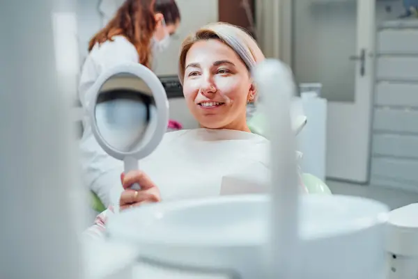 在口腔科诊所椅子上坐着的年轻微笑妇女的画像 并在牙齿美白后看着镜子检查她的牙齿 医疗保健和医疗保健行业的概念形象 — 图库照片