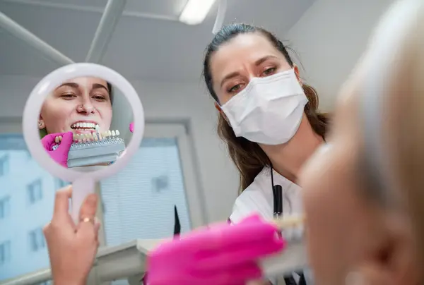 年轻的女性坐在口腔科诊所的椅子上 微笑着看着镜子露出她的牙齿 选择带有阴影标签的牙齿颜色 医疗保健 口腔卫生和医疗行业概念形象 — 图库照片