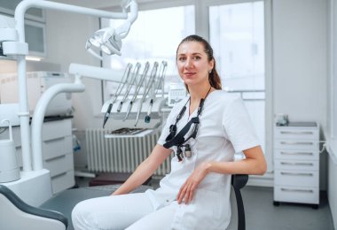 Modern diş sağlığı kliniğinde stomatoloji kürsüsünün yanında oturan beyaz önlüklü, gülümseyen genç dişçi kadın portresi. Sağlık ve sağlık sektörü konsepti.
