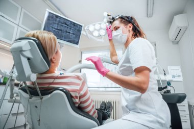 Dişçi kliniğindeki hasta modern sağlık koğuşunu ziyaret ediyor. Dişçi kadın doktor üniforma giyip büyüteç takmış ve ışığı işaret ediyor. Hasta sandalyede oturuyor. Sağlık ve sağlık sektörü konsepti