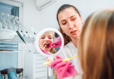 Dişçi doktor stomatoloji kliniğinde diş fırçasıyla diş hijyenik prosedürleri yaparken küçük kız aynaya bakıyor. Sağlık, çocuk sağlığı ve sağlık sektörü konsepti.