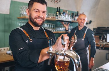 Siyah üniformalı şık sakallı barmen kameraya gülümsüyor, bar tezgahına bira vuruyor ve garsonu tepsiyle dürtüyor. Başarılı insanlar takım çalışması, arkadaşlık, demleme ve restoran endüstrisi konsepti imajı