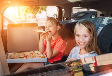 İki pozitif gülümseyen kız kardeş, arabanın arka koltuğunda çocuk koltuklarında pişmiş İtalyan pizzası yemekten mutlu olurlar. Mutlu çocukluk, fast food yemek, ya da otomobil yolculuğu öğle yemeği tatili konsepti.