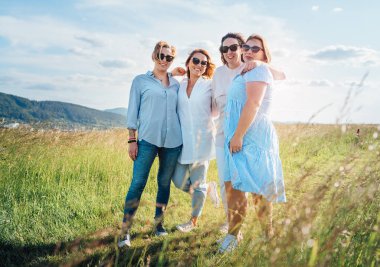 Yüksek yeşil çimen tepesinde yürürken kucaklaşan ve gülümseyen dört kadının portresi. Kameraya bakıyorlar. Kadın arkadaşlığı, ilişkiler ve mutluluk kavramı imajı.